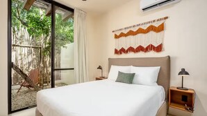 Een slaapkamer met een bed, twee nachtkastjes, een airconditioningapparaat, een raam met uitzicht op groen en een macramé wanddecoratie.