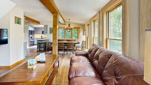 A spacious living area with a leather sofa, wooden flooring, a dining table, and large windows offering natural light. - NEW Cranmore Basecamp Ski/Downtown/Central (Conway)