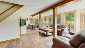 A spacious living area with a wooden floor, a sofa, a coffee table, and a dining table with chairs. Large windows provide natural light and views of the outdoors. - NEW Cranmore Basecamp Ski/Downtown/Central (Conway)