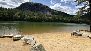 A sandy beach with large rocks and a calm lake surrounded by dense forest and a mountain. - NEW Cranmore Basecamp Ski/Downtown/Central (Conway)