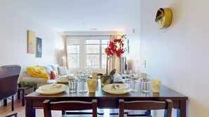 A dining area with a table set for four, a sofa with a yellow blanket, and a large window with curtains. - Modern Comforts ,Free Gym Access (Stamford)