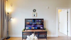 A living room with a flat-screen TV on a stand, a floor lamp, a small table with a plant, and a door to the right. - Modern Comforts ,Free Gym Access (Stamford)