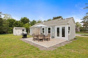 Ein eingeschossiges Haus mit einer Terrasse, die einen Tisch, Stühle, einen Grill und eine überdachte Sitzgelegenheit bietet.