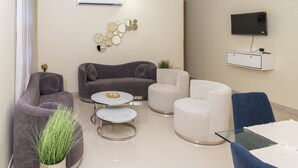 A modern living room with a grey sofa, a white armchair, a glass coffee table, and a flat-screen TV mounted on the wall.