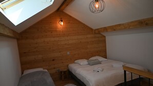 Ein gemütliches Schlafzimmer im Dachgeschoss mit schrägen Wänden, Holztäfelung, einem Oberlicht, einem Bett mit weißer Bettwäsche, einem kleinen Hocker und einer Pendelleuchte.