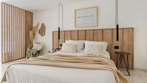 A modern bedroom with a wooden headboard, a large bed with white linens, a green throw blanket, and a minimalist decor including a framed picture and a small table.