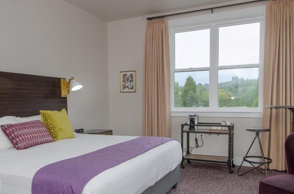 A hotel room with a bed, a desk, a chair, a window with curtains, and a picture on the wall.