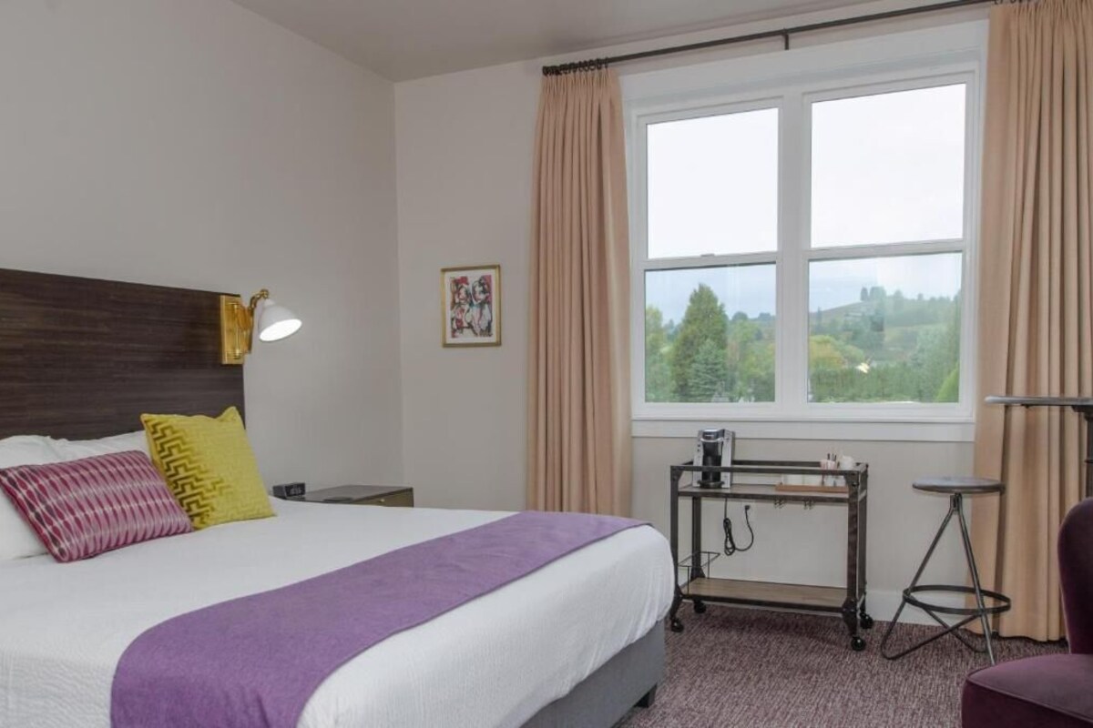A hotel room with a bed, a desk, a chair, a window with curtains, and a picture on the wall.