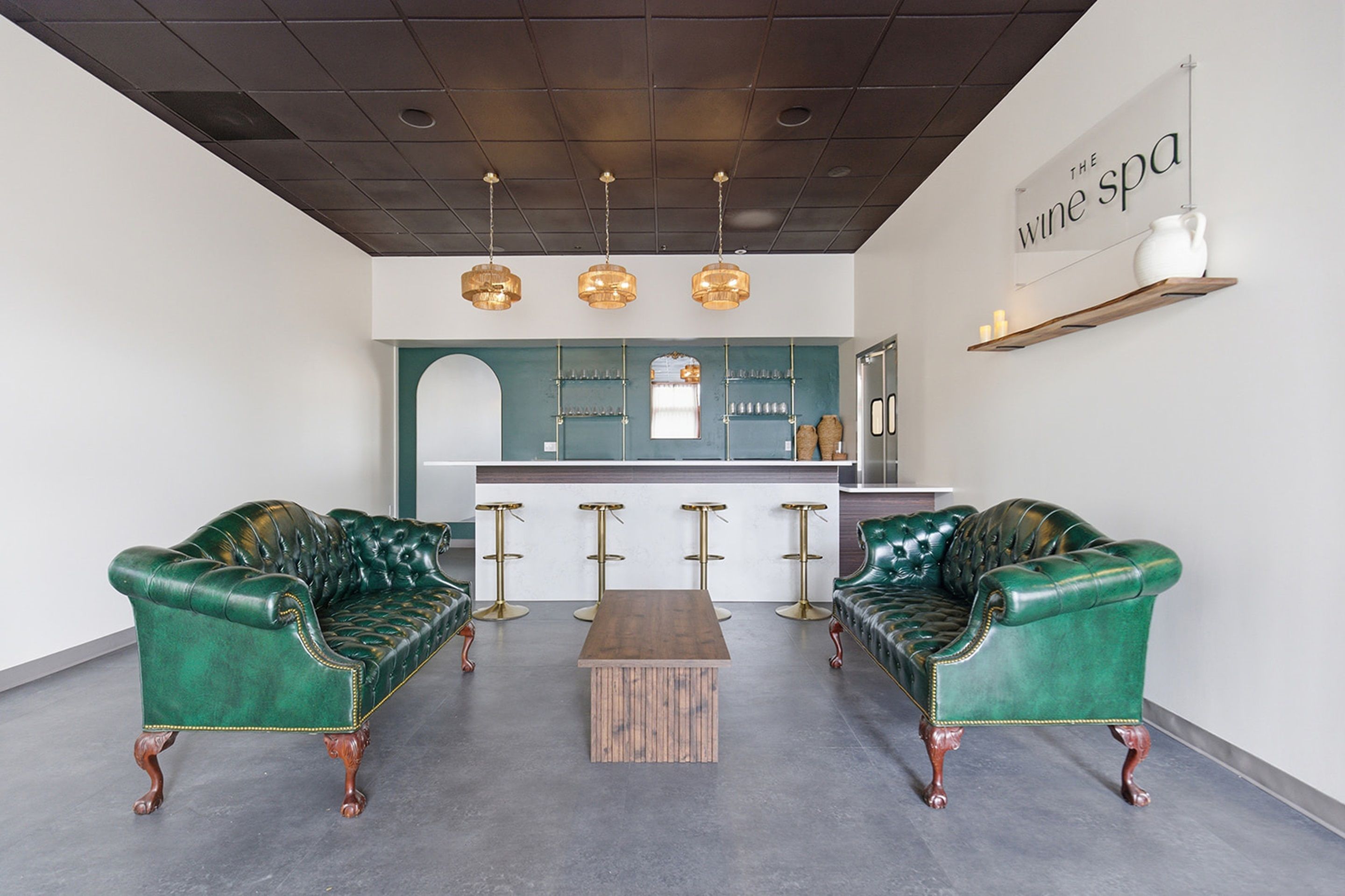A modern lounge area with two green leather sofas, a wooden coffee table, and a bar area with a teal accent wall.
