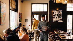 Interior de restaurante com mesas e cadeiras de madeira, janelas amplas e obras de arte nas paredes.