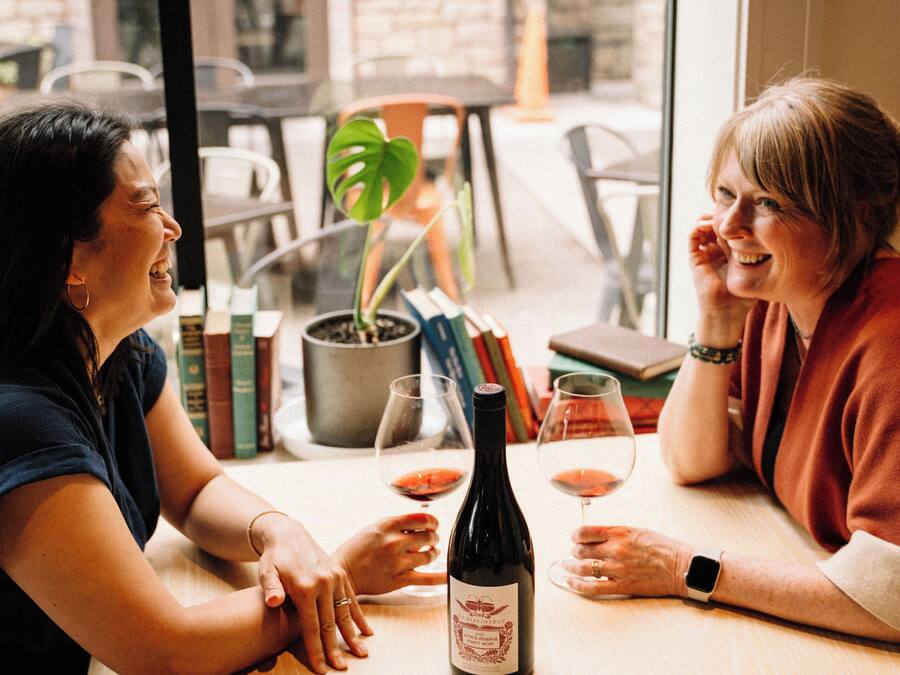 Two people sitting at a table with a bottle of wine and glasses, a potted plant, and books in a cafe setting.