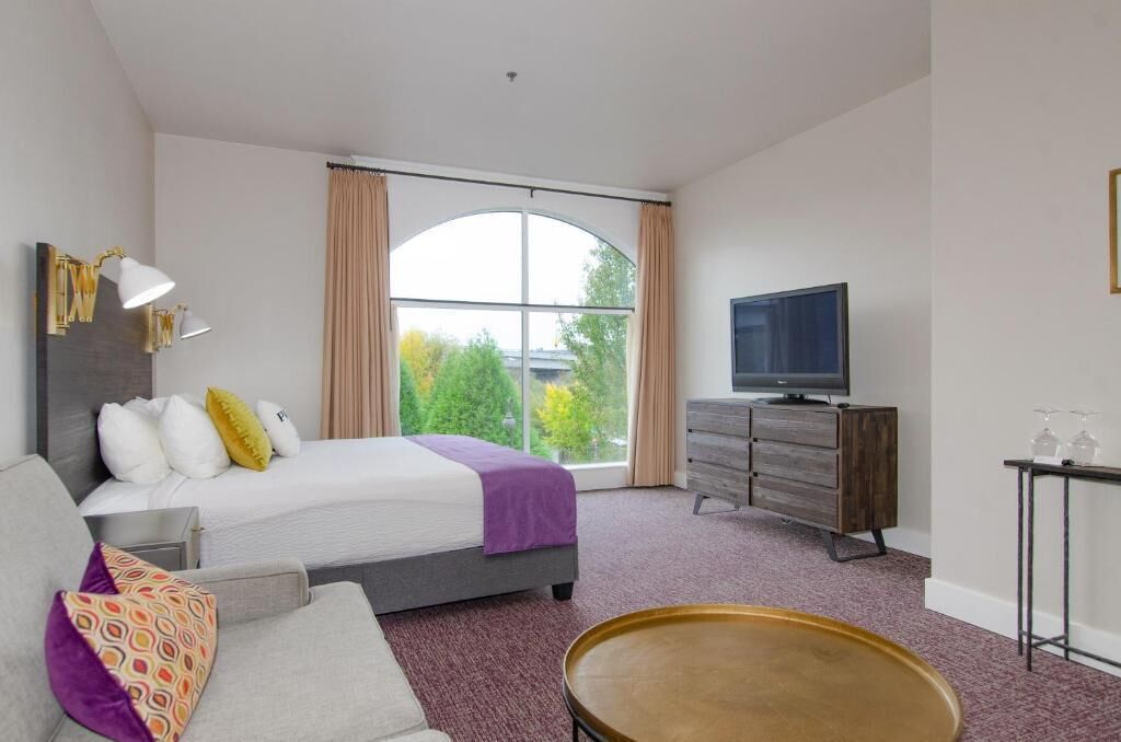 Habitación de hotel con cama, televisión, cómoda y un amplio ventanal con cortinas.