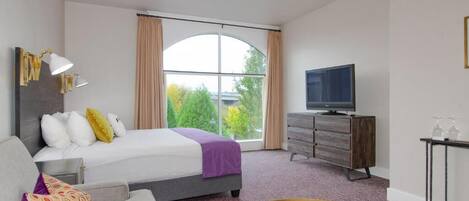 Habitación de hotel con cama, televisión, cómoda y un amplio ventanal con cortinas.