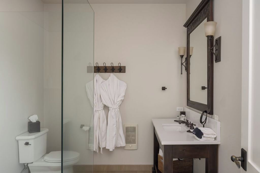 A bathroom with a glass shower enclosure, a white toilet, a sink with a mirror, and two white robes hanging on a rack.