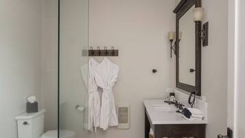 A bathroom with a glass shower enclosure, a white toilet, a sink with a mirror, and two white robes hanging on a rack.