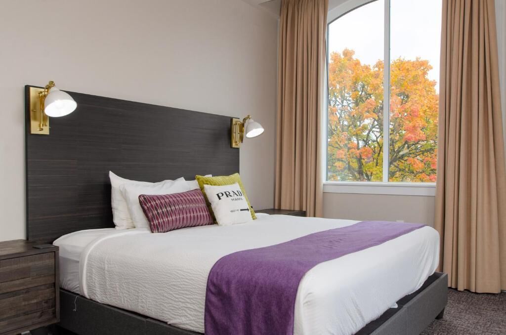 Ein ordentlich gemachtes Bett mit weißer Bettwäsche und einer violetten Decke, zwei Nachttischlampen und ein Fenster mit Vorhängen.
