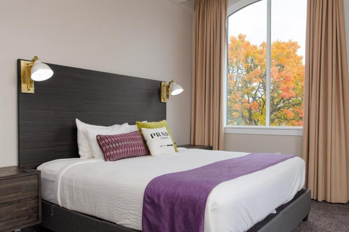 A neatly made bed with white bedding and a purple throw, two bedside lamps, and a window with curtains.