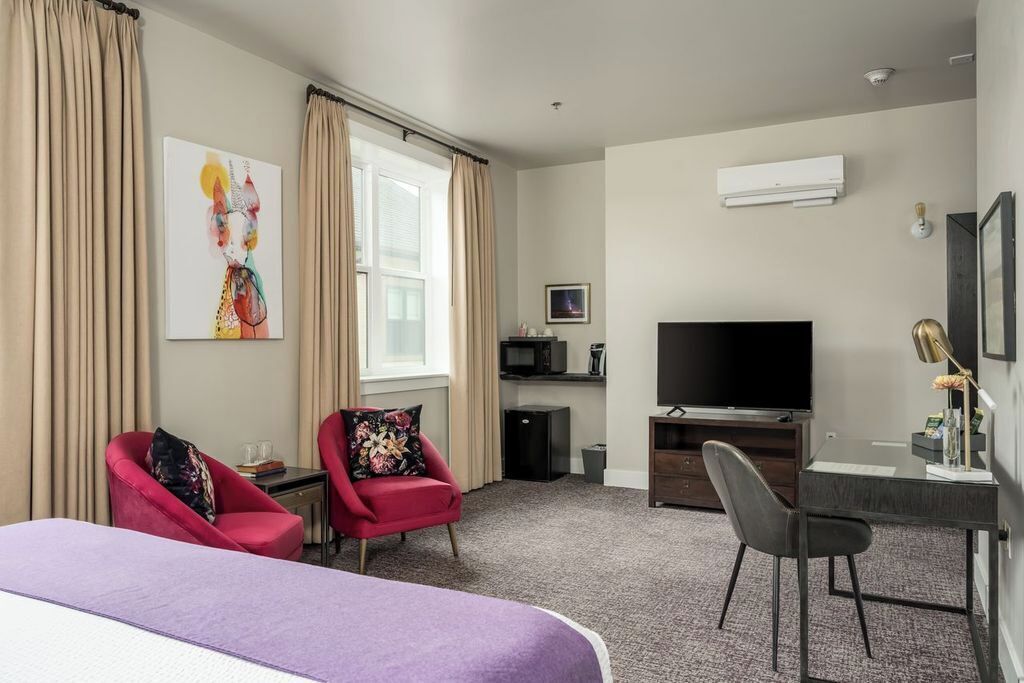 A hotel room with a bed, two red armchairs, a desk with a chair, a television, and an air conditioning unit.