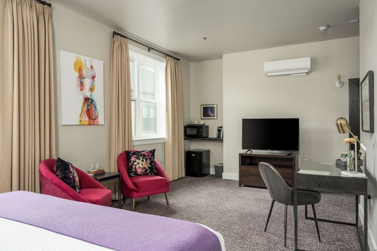 A hotel room with a bed, two red armchairs, a desk with a chair, a television, and an air conditioning unit.
