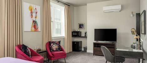 A hotel room with a bed, two red armchairs, a desk with a chair, a television, and an air conditioning unit.