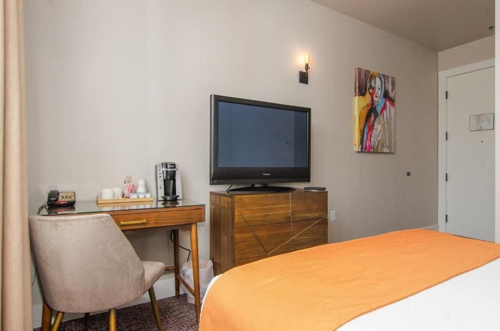 Habitación de hotel con una cama, un televisor sobre un mueble de madera, un escritorio con una silla y un cuadro en la pared.