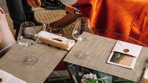 Um homem serve vinho em uma taça à mesa, com uma mulher sentada do outro lado.