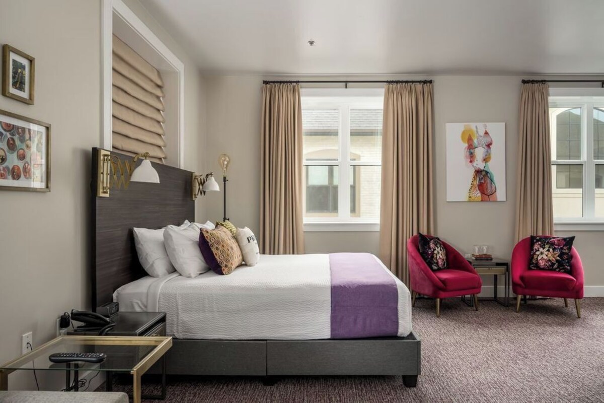 A hotel room with a bed, bedside table, lamp, chair, and artwork on the wall.