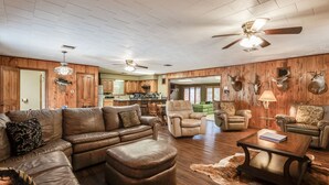 A spacious living room with a sectional sofa, leather armchairs, and a coffee table. The room features wooden paneling, mounted deer heads, and a ceiling fan.