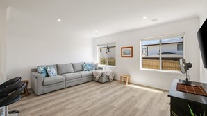 Een moderne woonkamer met een grijze modulaire bank, een klein houten tafeltje en een zwarte stoel. Twee ramen bieden uitzicht op een houten schutting en een gebouw.