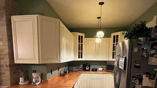 Una cocina con suelo de madera, armarios blancos, una isla integrada, un refrigerador y un colgante de luz.
