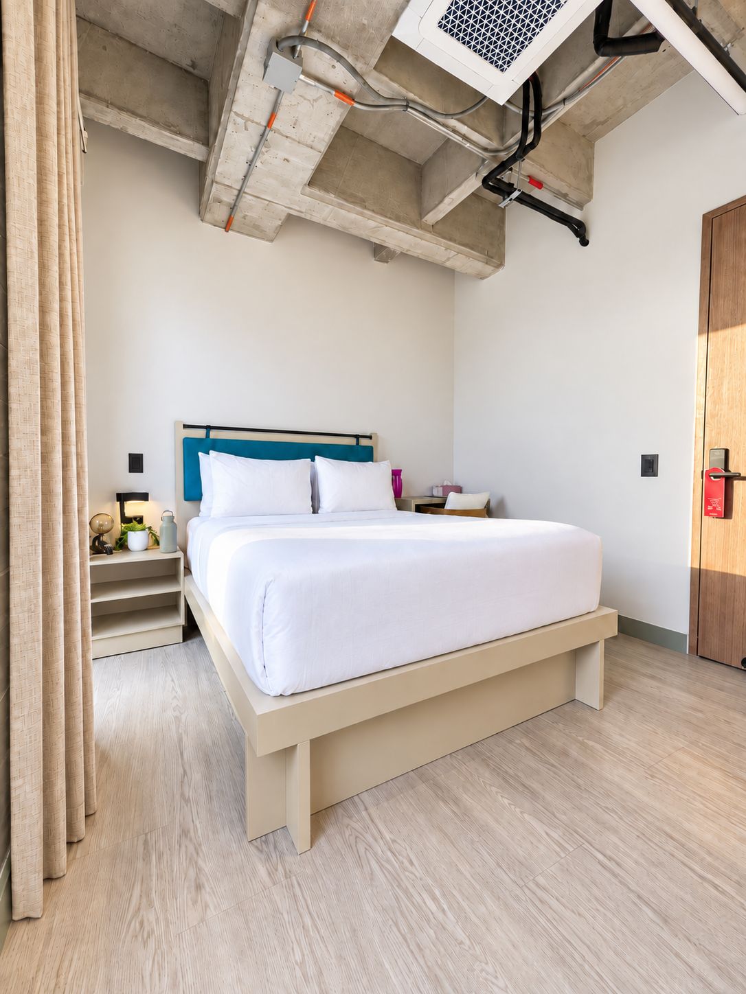 A modern hotel room with a large bed, wooden headboard, bedside tables, and a visible ceiling with exposed pipes and ducts.