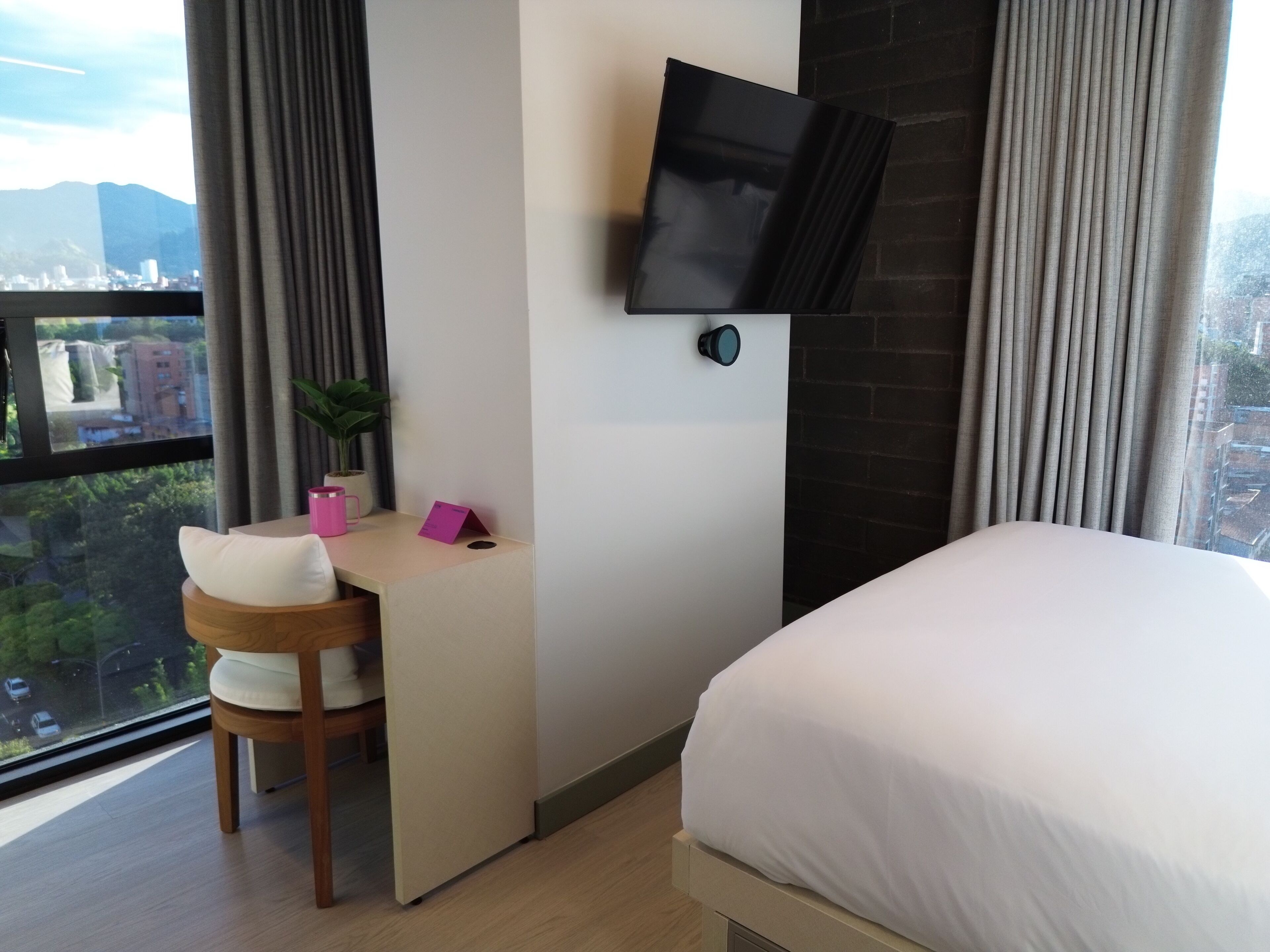 A hotel room with a bed, a desk with a chair, a television mounted on the wall, and a view of the cityscape through the window.
