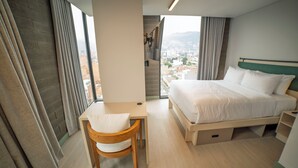 Une chambre d’hôtel équipée d’un lit, d’un bureau, d’une chaise et offrant une vue sur le paysage urbain grâce à de grandes fenêtres.