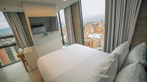A hotel room with a bed, a TV, a small table, and a view of a cityscape through large windows. - CITIO Hotel - Laureles (Medellín)