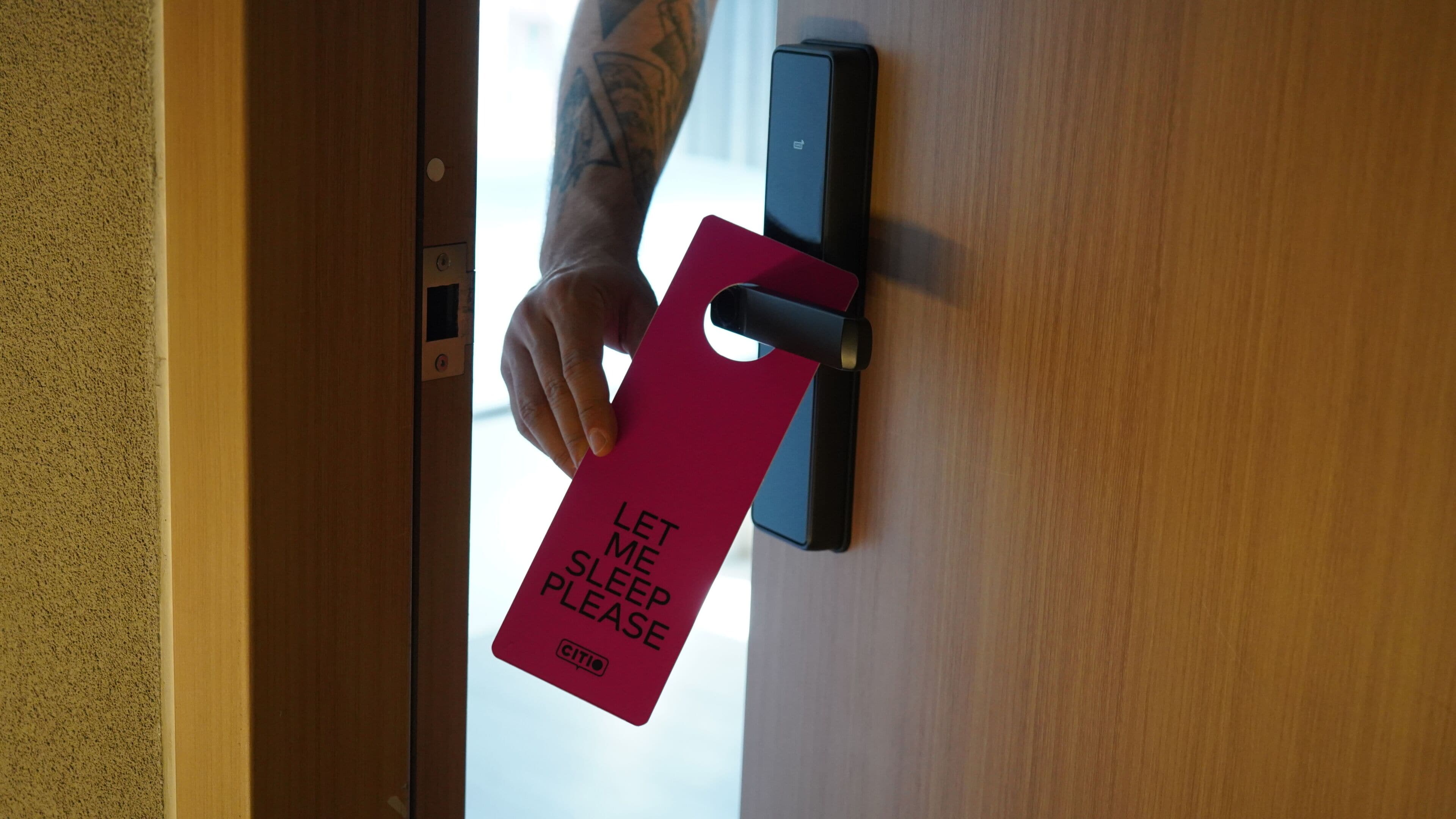 Eine Hand hält eine rote Karte mit dem Aufdruck 'LET ME SLEEP PLEASE', die an einem Türschloss angebracht ist.
