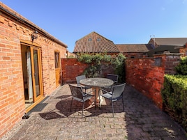 A brick building with a tiled roof, a patio with a round table and chairs, and a door leading to an interior space.