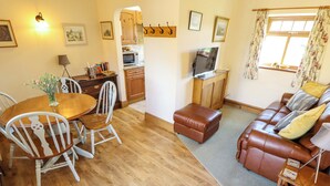 A living room with a brown leather sofa, a wooden dining table with chairs, a television on a wooden cabinet, and a kitchen area in the background. - Ticklepenny (Louth)