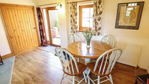 A dining area with a wooden table, white chairs, a vase with flowers, a window with floral curtains, and a framed picture on the wall. - Ticklepenny (Louth)