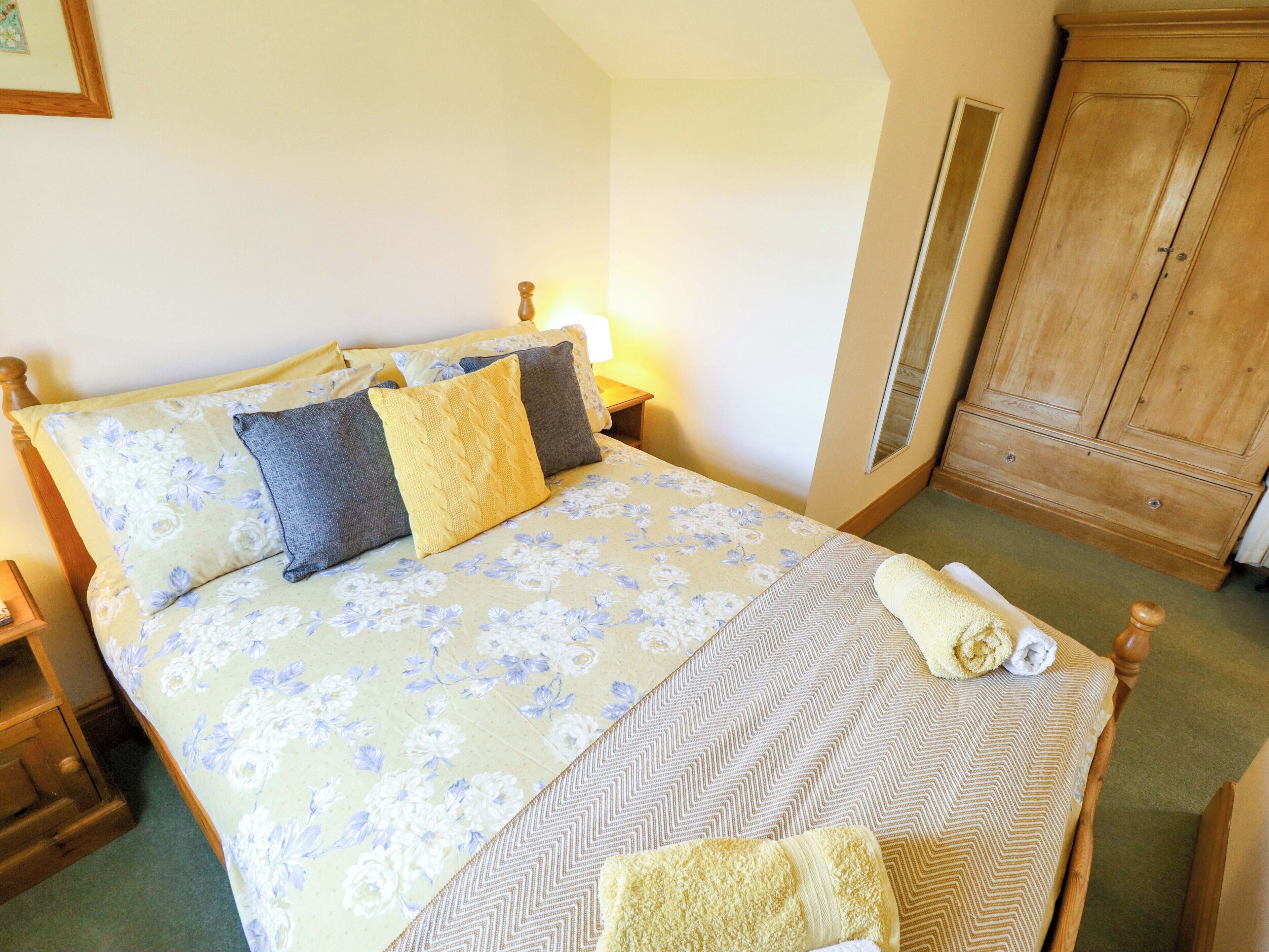 A neatly made bed with floral bedding, a wooden wardrobe, and a nightstand with a lamp.