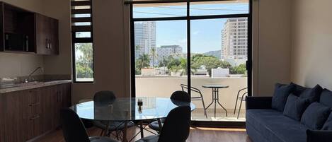 A modern living room with a glass dining table, dark sofas, and a large window offering a city view.