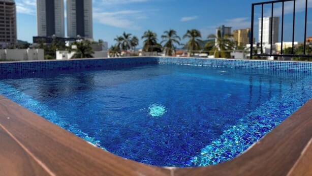 A swimming pool with a wooden deck edge, surrounded by palm trees and tall buildings in the background. - Depa A few blocks from the Beach and Golden Zone ByGPS (Mazatlán)