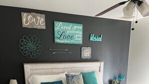 A bedroom with a wooden bed, white bedding, teal and blue pillows, a nightstand with a lamp, and wall decorations including a 'love' sign and a 'loved you' sign. - Spacious 4-bedroom house, perfect for entertaining in the heart of Ridgecrest. (Ridgecrest)