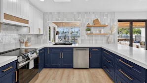 Une cuisine moderne avec des meubles blancs, des touches de bleu et une vue sur l’espace extérieur aménagé pour se détendre.