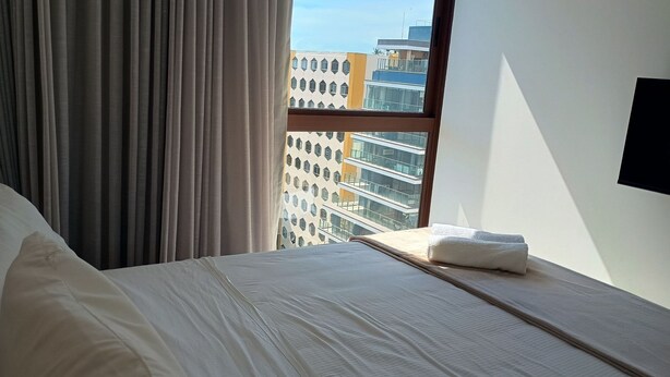 A hotel room with a bed, a window with curtains, and a view of a building with multiple windows. - Cozy Apt Sea View Barra - Nau0810 (Salvador)