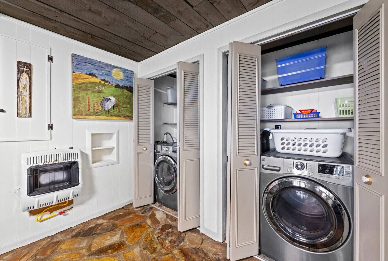 A laundry room with a stone floor, a washing machine, a dryer, and a shelf with laundry baskets.