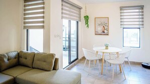 Un salon comprenant un canapé d’angle, une table à manger avec des chaises et de grandes fenêtres équipées de stores.
