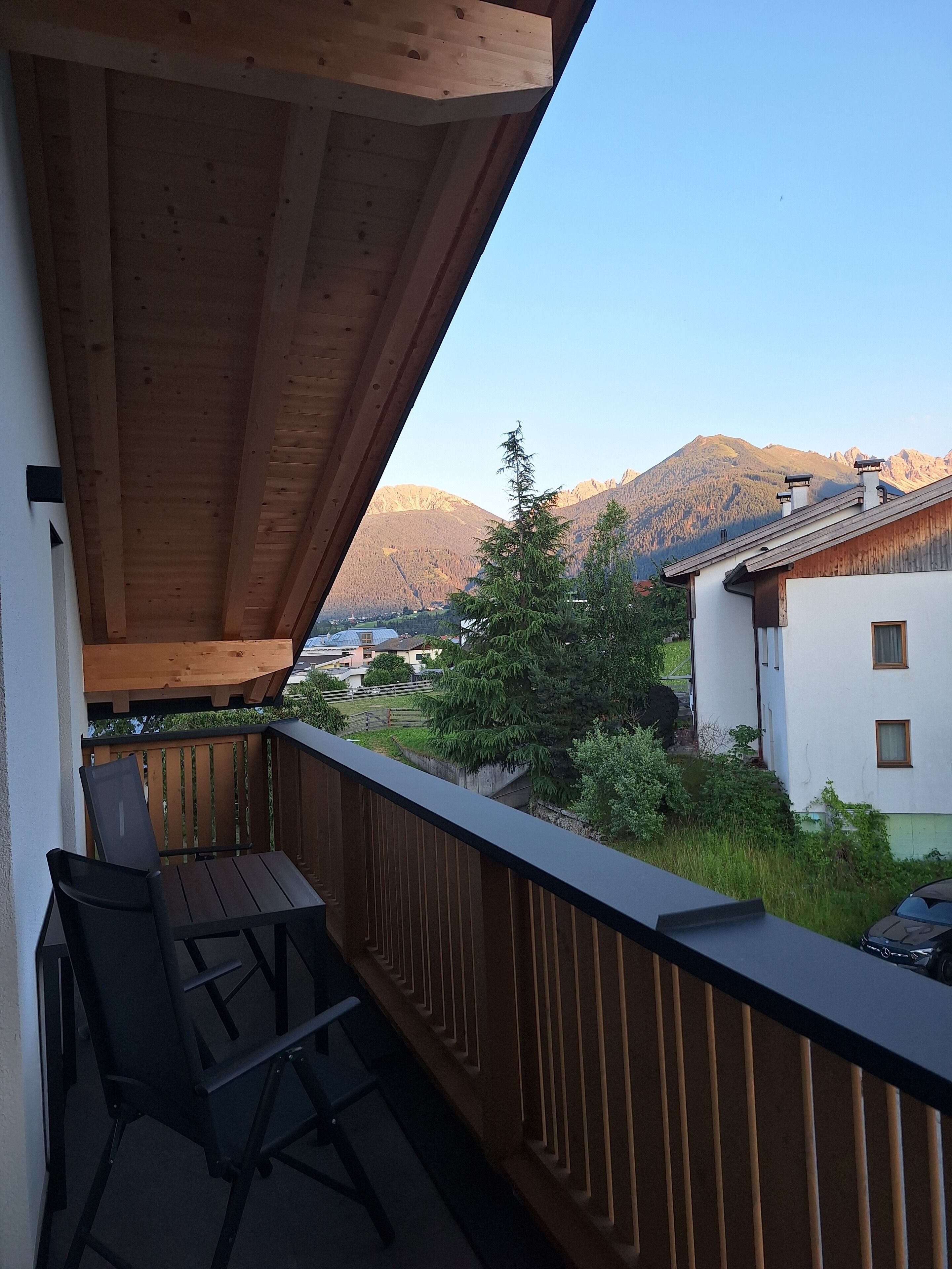 Een balkon met houten plafond, een tafel en twee stoelen. Achtergrond met bergen en uitzicht op een dorp.
