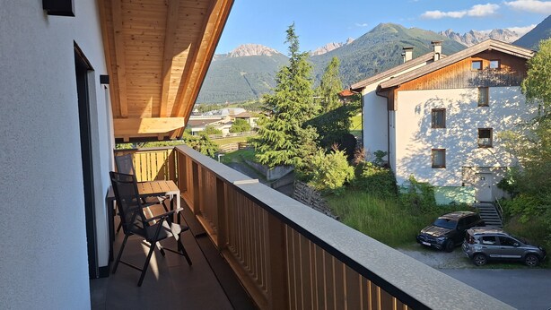 Un balcon avec des balustrades en bois, une table et deux chaises. En arrière-plan, on aperçoit des bâtiments, une montagne et un ciel dégagé.