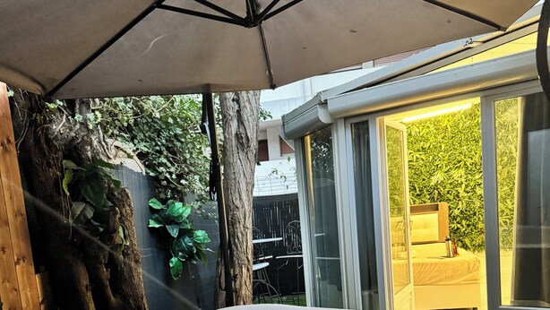 Un jacuzzi inflable en un patio trasero con un porche cubierto, un tronco de árbol de madera y un ventanal corredizo que da a un patio con mesa y sillas.