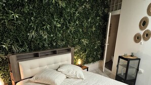 Un dormitorio con una cama grande, una pared decorativa con plantas, una mesita de noche con lámpara y una pequeña mesita con un adorno.